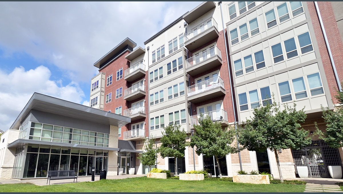 A modern multi-story apartment building with a mix of brick and glass exteriors. The building features balconies, large windows, and is surrounded by a well-maintained lawn area.