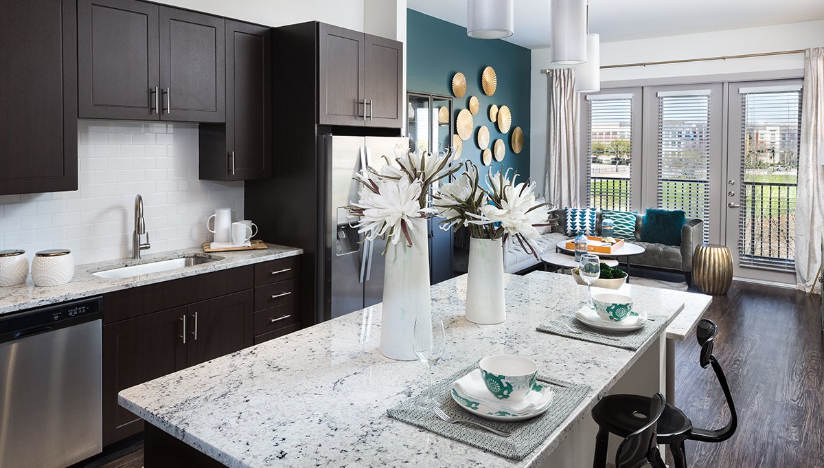 A model kitchen at Crest at Las Colinas Station. The kitchen island has place settings ready across from the dark cabinets and stainless steel appliances. A dining table sits along the windows in the background, the courtyard seen just outside.