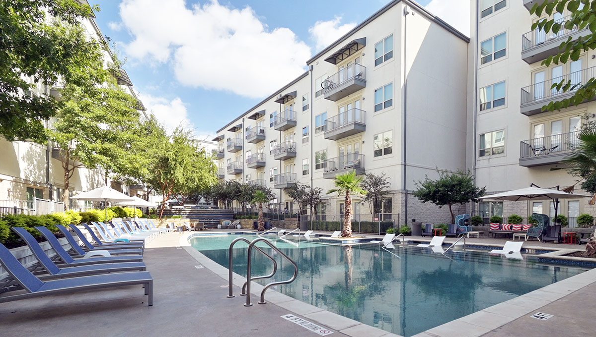 A modern apartment complex surrounds a rectangular outdoor swimming pool with lounge chairs and umbrellas on the deck.