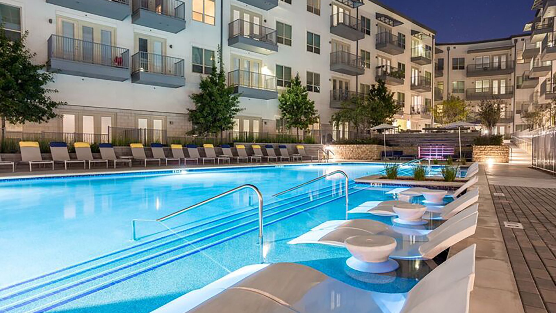The outdoor pool at Crest at Las Colinas Station as seen from the tanning shelf at night. The apartment building surrounds the pool with green shrubs intermixed.