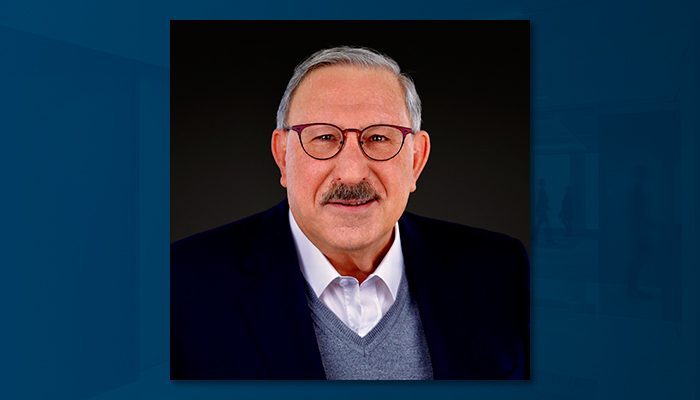 Professional portrait of Ed Polich, a man with short gray hair, glasses, and a mustache, wearing a dark blazer, white shirt, and gray sweater vest, smiling against a dark background.