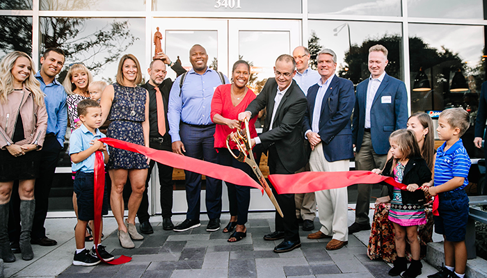 A group of people stand outside the front door of a new retail space and happily cut a large, red ribbon with gold scissors to celebrate a grand opening.