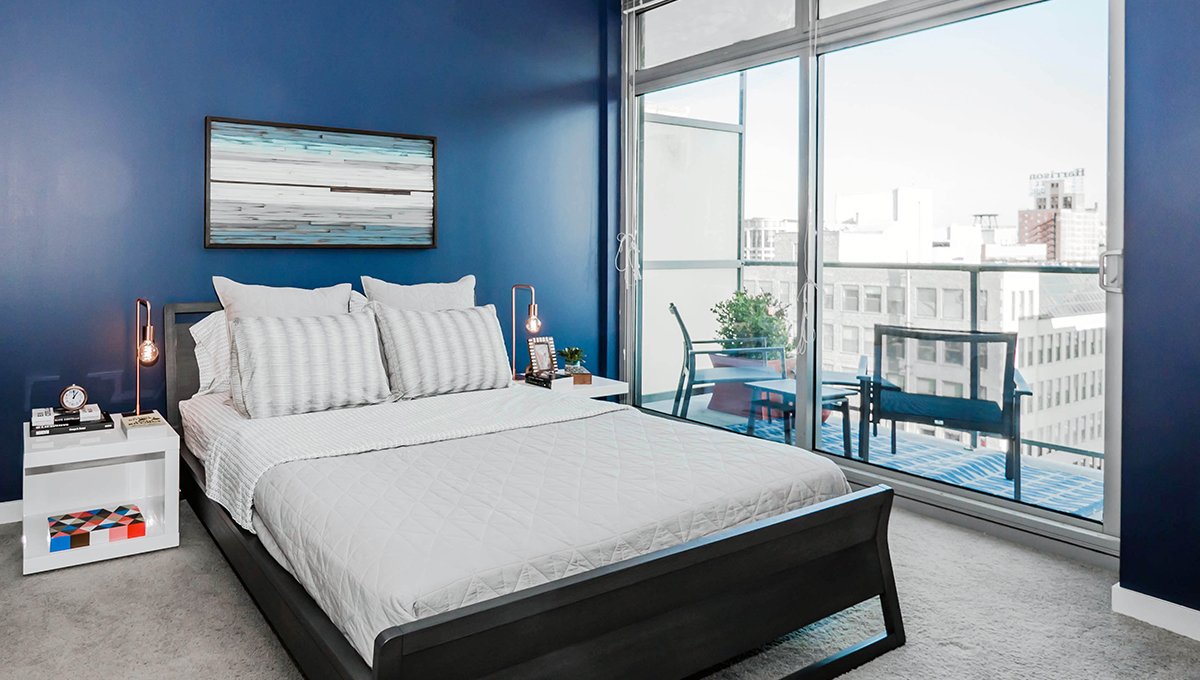 A queen sized bed sits a room with blue walls. A balcony is seen just out the window with two lounge chairs on it. The city is seen in the distance.