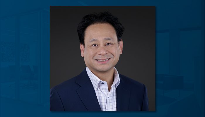 Professional portrait of Blas Puzon, a man with short black hair, wearing a navy blazer and a white checkered shirt, smiling against a dark background.