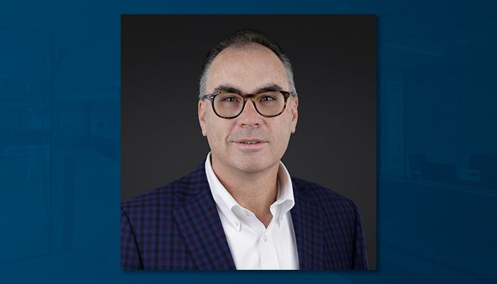 Professional portrait of James Hayes, a man with short gray hair, glasses, and a clean-shaven face, wearing a white shirt and a navy checkered blazer, against a dark background.