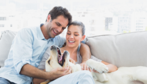 A couple sitting on a white couch cuddling their dog.