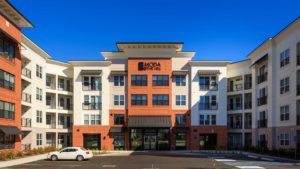 Looking straight on at the exterior front entrance to Moda at The Hill. The façade is a mix of red brick, grey and white. The sky is blue. There is only one white car parked in the lot in front.