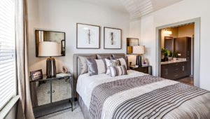 A typical bedroom at North 680. A queen sized bed has mirrored end tables and table lamps on each side. The dark cabinets of the bathroom can be seen through a doorway in the background.