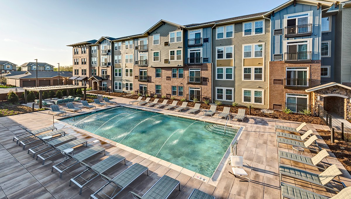 An elevated view of the outdoor pool at North 680. Lounge chairs surround the pool with water jets spraying arcs across the pool. The apartment building is in the background.