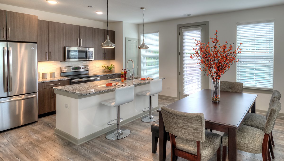 A kitchen and dining room in one of the townhomes at North 680. A dining table site to the right with a bright red flower centerpiece. The dark wood cabinets and stainless steel appliances of the kitchen are in the background to the right behind the kitchen island.