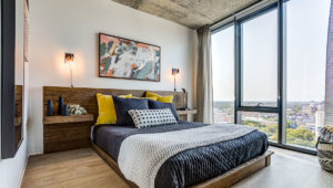 A bed with wooden headboard and end tables sits next to floor-to-ceiling windows, the city seen outside.
