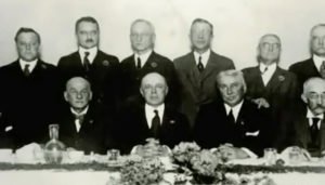 Two rows of gentlemen in black ties, seated and standing behind, at a banquet table.