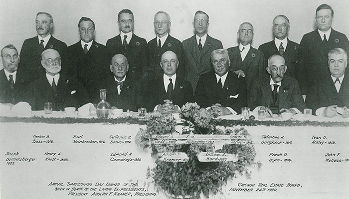 Two rows of gentlemen dressed in black ties posing for a photograph at a dinner table, black and white photo, circa 1920.