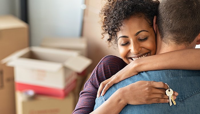 A woman hugs a man holding keys to their new home, moving boxes in the background.