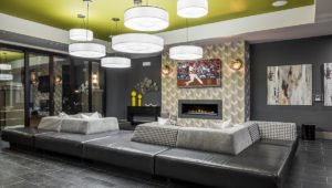 A large black sofa sits before a fireplace in the resident lounge at EVO Living. Several circular lamps hang from the vividly painted green ceiling. A flat screen television on the wall shows a baseball game playing.
