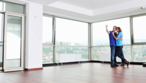 A couple happily looks at their new apartment. It is currently empty and there are floor to ceiling windows with a view of a city.
