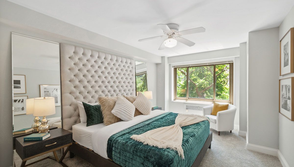 A modern bedroom with a tufted headboard, green bedding, and decorative pillows. A window illuminates the room, and a white armchair sits beside a small table with lamps.