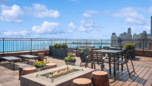 Rooftop patio with seating, a glass-encased fire pit, plants, and city skyline in the background under a blue sky.