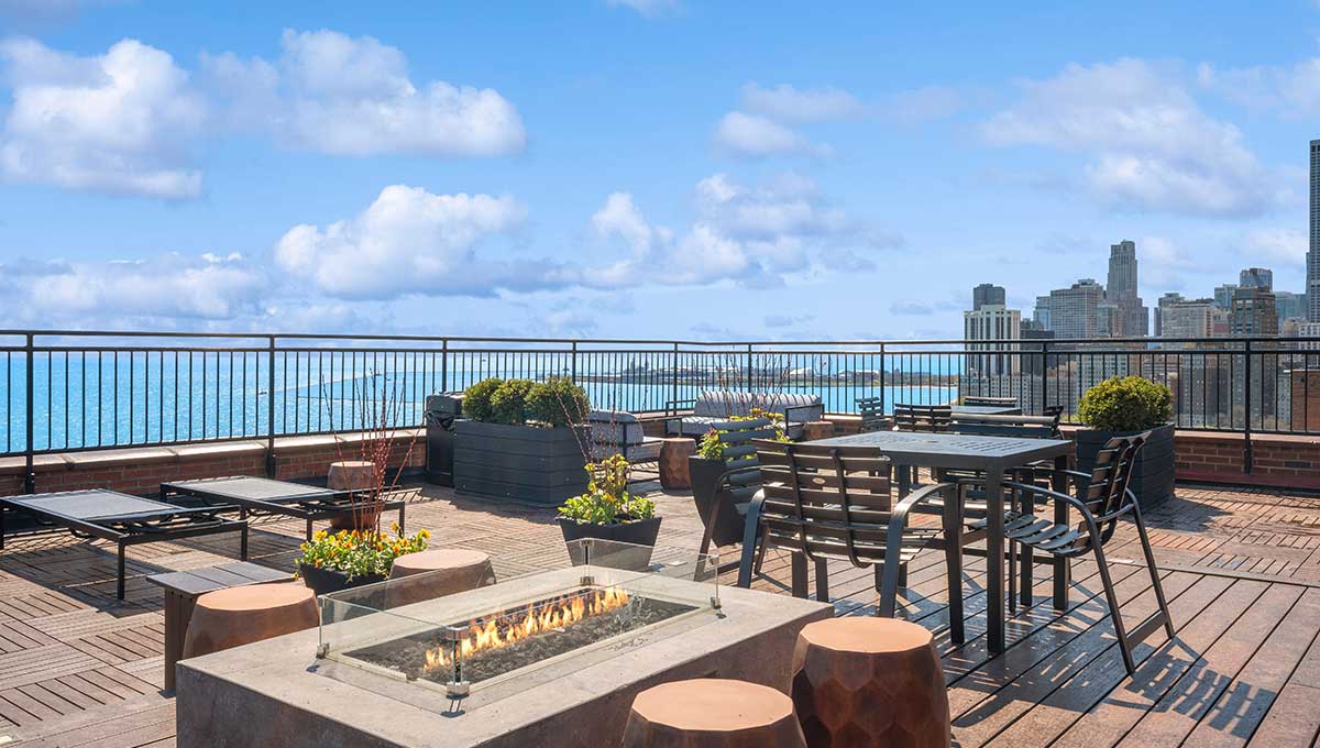 Rooftop patio with seating, a glass-encased fire pit, plants, and city skyline in the background under a blue sky.