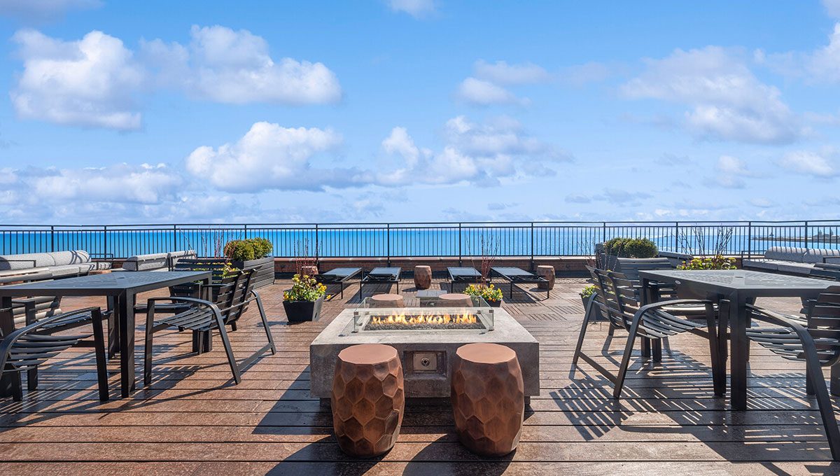 Rooftop patio with ocean view, featuring tables, chairs, a central fire pit, and potted plants under a partly cloudy sky.