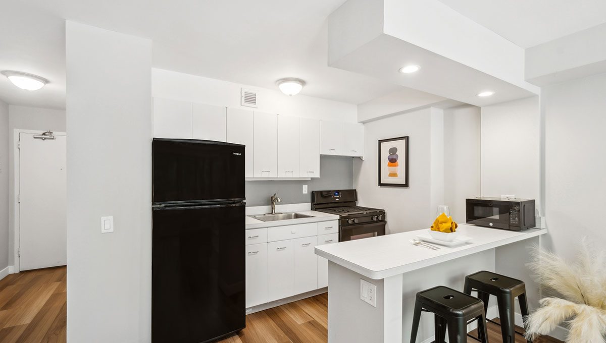 Modern kitchen with white cabinetry, black fridge, and stove. A small island with two black stools, a microwave, and decor complete the space. Wooden flooring and white walls are present.