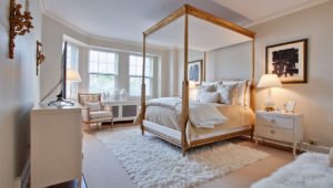 A bedroom at 1420 North Lake Shore with a canopy bed over a lush rug. A white dresser is along the left wall and a club chair sits beneath the windows.