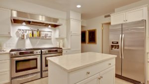 A kitchen at 1420 North Lake Shore with all white cabinets and stainless steel appliances. A kitchen island is in front with the a double oven and stove stop behind to the left and large refrigerator to the right.
