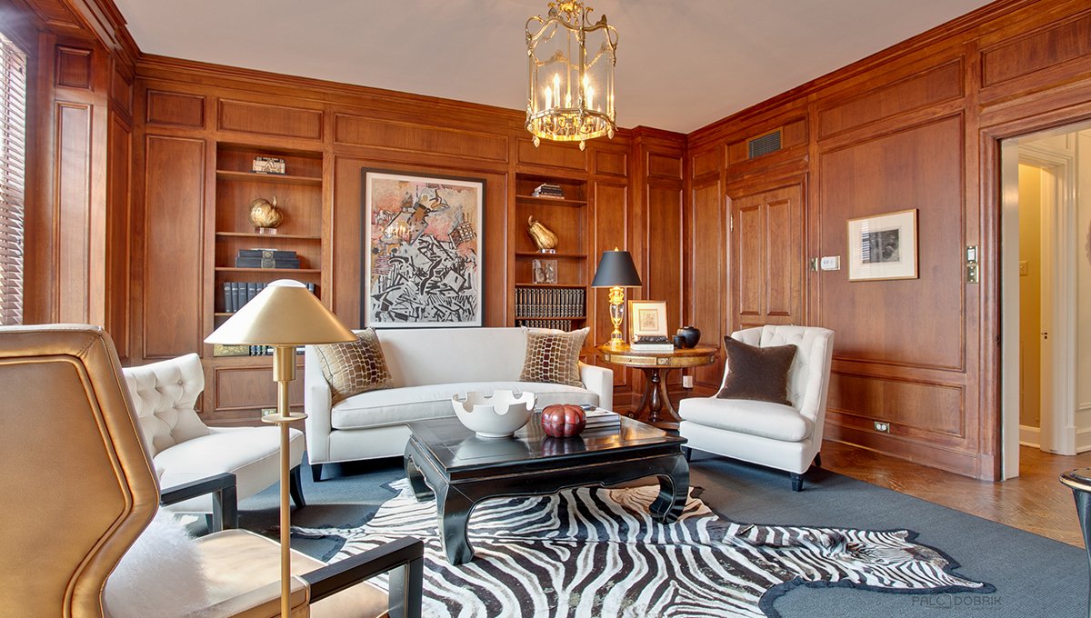 A living room at 1420 North Lake Shore with a white couch and matching club chairs around a coffee table over a zebra-print rug. The walls are wood panels with built-in bookshelves.