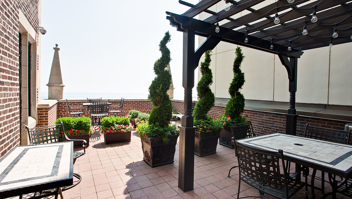 A rooftop terrace at 1420 North Lake Shore with several cafe tables and chairs. Various potted plants decorate the area and Lake Michigan is seen in the distance.