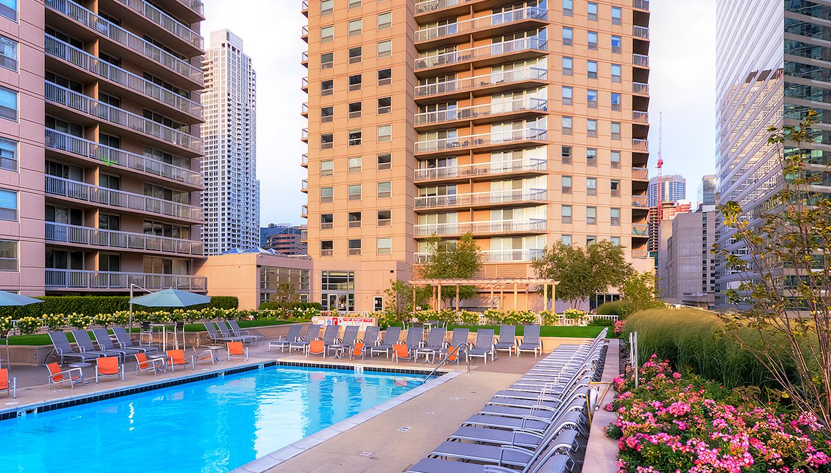 Looking across the outdoor pool and sundeck at the apartment tower of Grand Plaza. The pool is surrounded by patio chairs and greenery. The tower is rises up into the skyline with balconies on each floor.