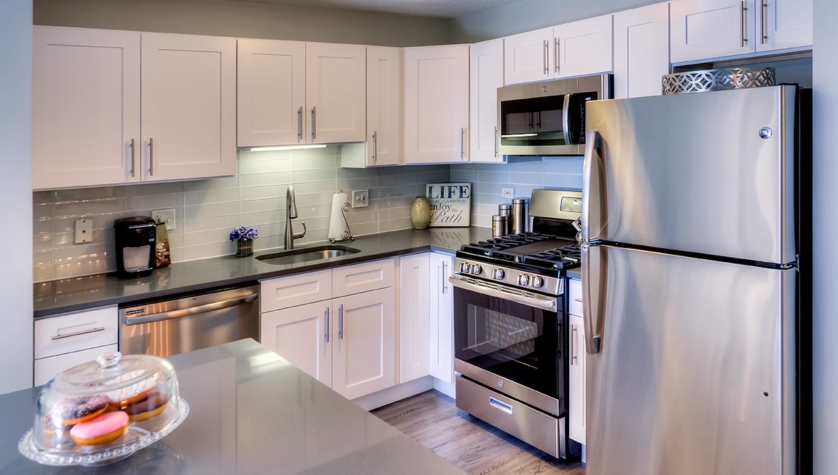 A kitchen in an apartment at Grand Plaza featuring white cabinets and grey countertops with stainless appliances. A glass cake stand is on the kitchen island with very delicious-looking donuts.