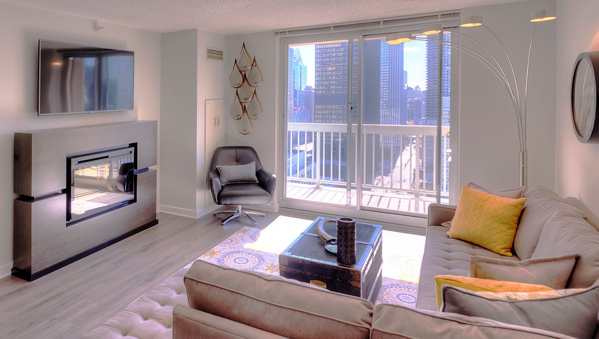 A living room at Grand Plaza apartments with a sectional couch and lamp on the right and a built-in fireplace with flat screen above across on the left. The city is seen outside the sliding glass door and balcony in the middle.