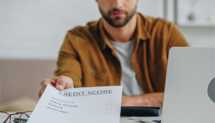 A bearded man sits before a laptop and hands a sheet towards the camera that has his credit score checked as "Excellent".