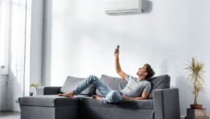 A man lays on his couch trying to stay cool by clicking the remote to the air conditioning unit above him.