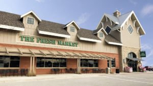 An exterior view of The Fresh Market at Lincolnshire Marketplace with a red brick base and beige siding. An awning covers seating beneath the retail windows.