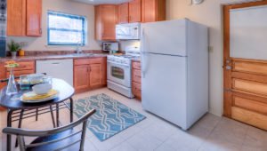 A kitchen in a residence at Homestead Apartments with honey oak cabinets and white appliances. A cafe table is on the left and a doorway on the right.