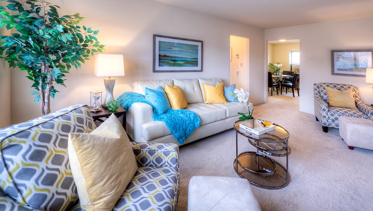 A living room in a unit at Homestead Apartments with large white, blue and yellow patterned chairs and couch set in the room. Doorways in the background lead to a hallway and the dining room.