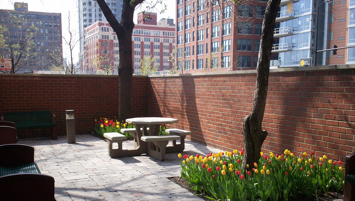 A red brick wall encloses the patio. A circular patio table and benches is surrounded by trees and red and yellow tulips. The city is seen on the other side of the wall.