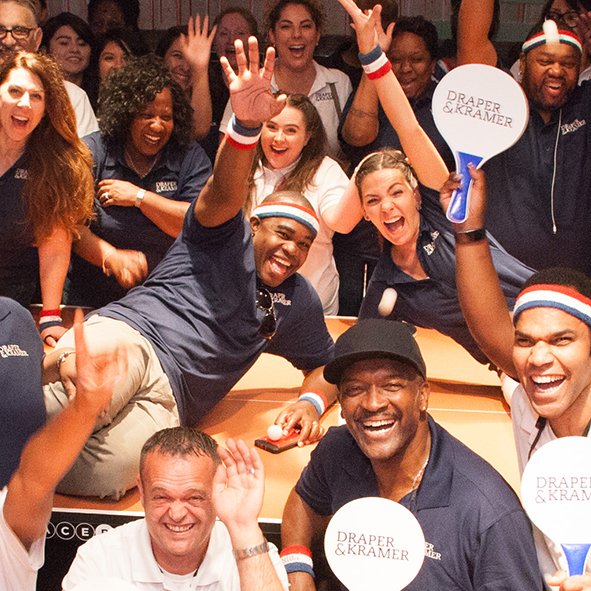 A group of Draper and Kramer employees waving to the camera at a ping pong event. Many are holding up paddles that have the Draper and Kramer logo on them.