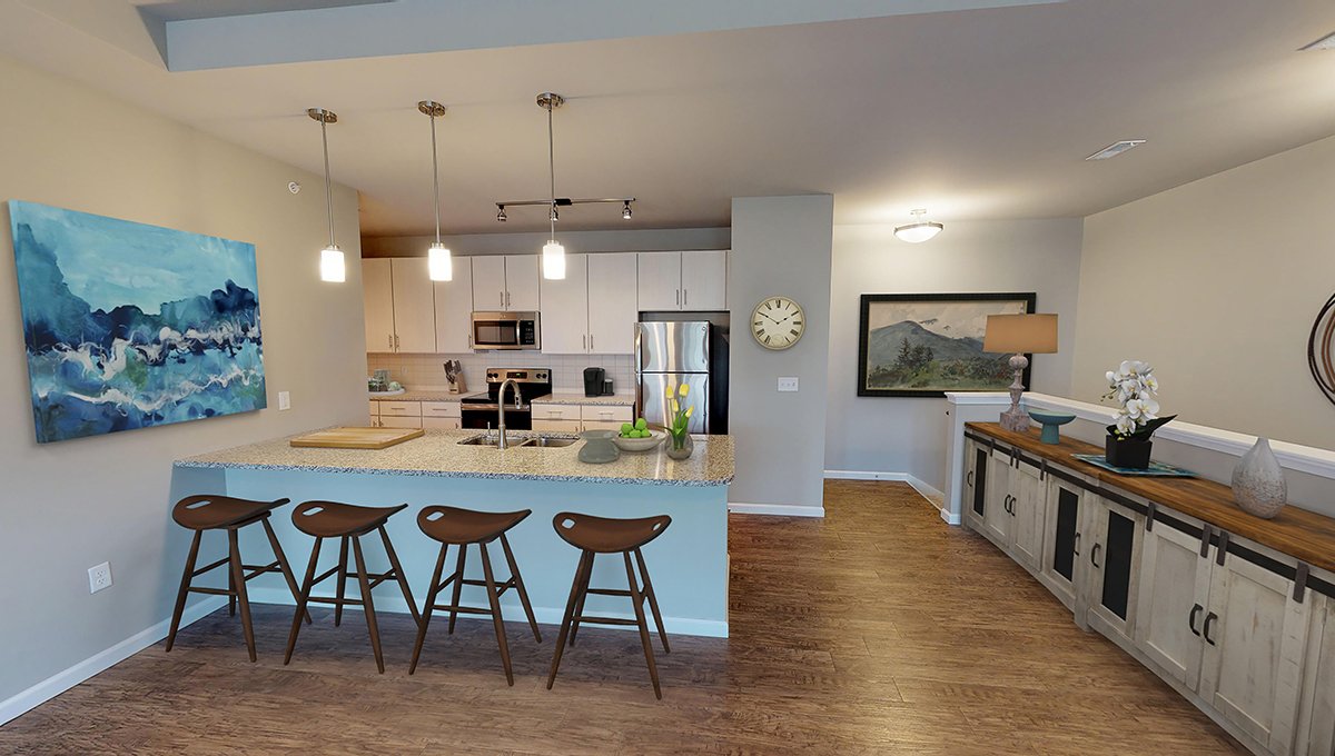 A virtually staged kitchen in an EVO Flats unit. The kitchen has white cabinets and stainless steel appliances. Four stools are placed at the kitchen counter with lighting above. To the right is the stairwell leading down and out of the unit.