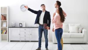 A couple is seen taking a tour of apartment with a leasing agent who is showing them a large room with furniture in the background.