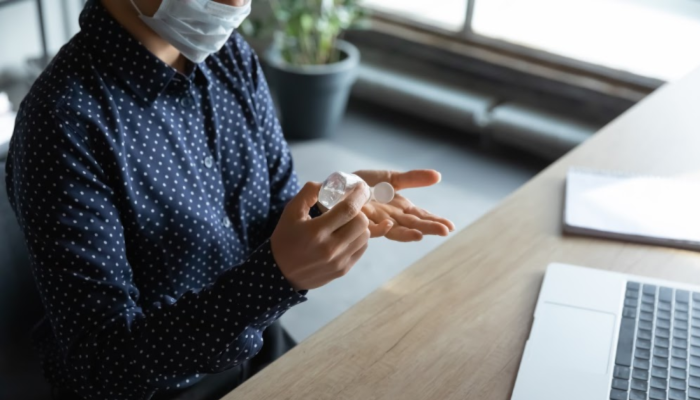 A man wear a medical face mask applies hand sanitizer while sitting at a desk.