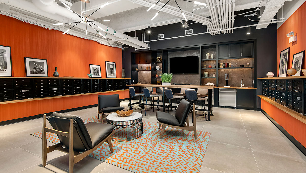 Looking in to the Clubroom at Wrigleyville Lofts. There are club chairs in a circle ahead with a high-top table behind. The far wall has a mounted television with beer taps beside. The side walls are orange with mailboxes built-in for residents.
