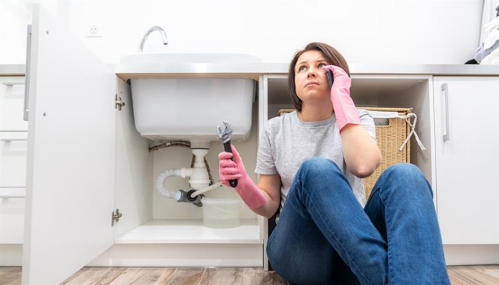 A woman sits under her sink holding a wrench in one hand and her cellular phone in the other.