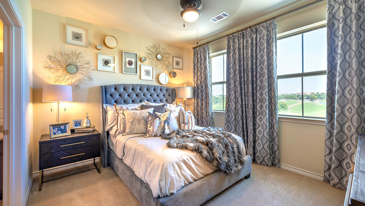 A bedroom within a residences at Adriatica Senior Living with bed and night stand before two large windows that show green field and clear sky outside.
