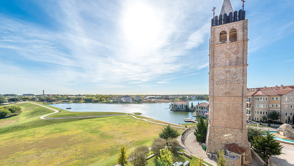 The Bell Tower at Adriatica Village sits before a large green field and a lake beyond that.