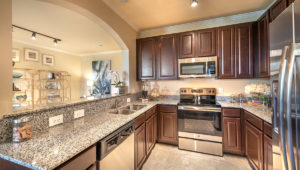A kitchen within a residence at Adriatica Senior Living with stone countertops, dark wood cabinets and stainless steel appliances. The wall is open to the living room along the left side.