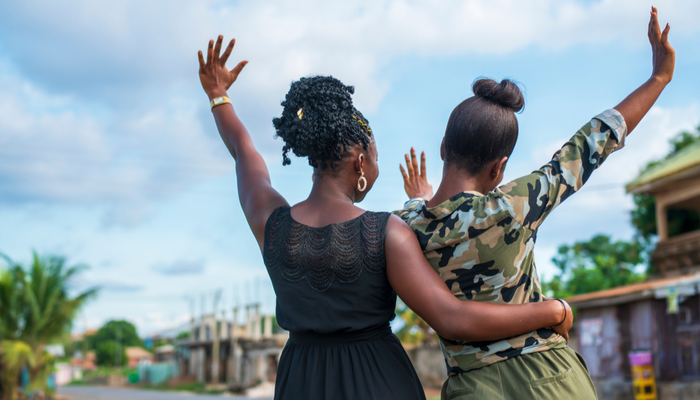 Two African-American women, arm in arm, wave happily on a sunny day.