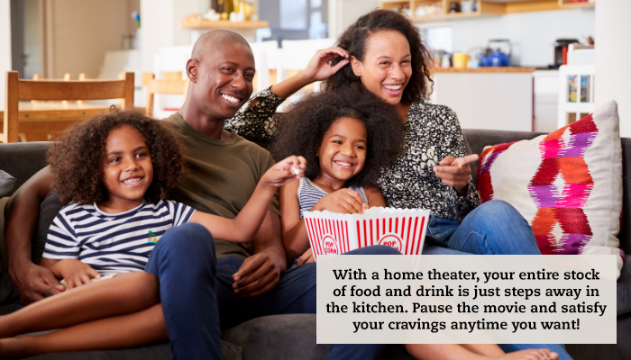 A family sits on a couch ready for movie night, complete with a large tub of popcorn. A caption reads, "With a home theater, your entire stock of food and drink is just steps away in the kitchen. Pause the movie and satisfy your cravings anytime you want!"