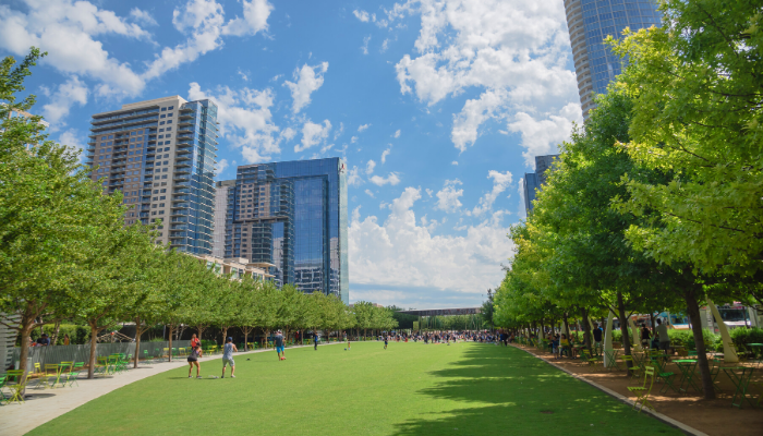 An open, grassy field in Dallas flanked by tall trees. Buildings are seen in the background on a partially cloudy day.